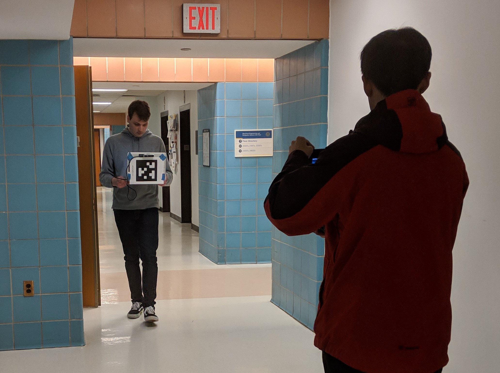 three men stand in a hallway.  Two are facing each other holding open laptops with April tags taped to the lids